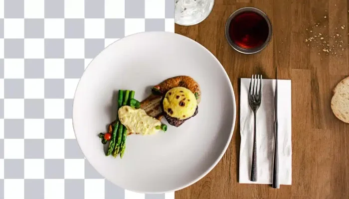 Em português de Portugal: Uma mesa de restaurante com um prato de comida. Atrás do prato, metade do fundo é um quadrado em tons de cinza e branco, como um arquivo PNG, e a outra metade são os elementos visíveis da mesa em madeira. Podem ser vistos meio copo de água, um copo de vinho, talheres, uma toalha, uma fatia de pão e migalhas de pão
